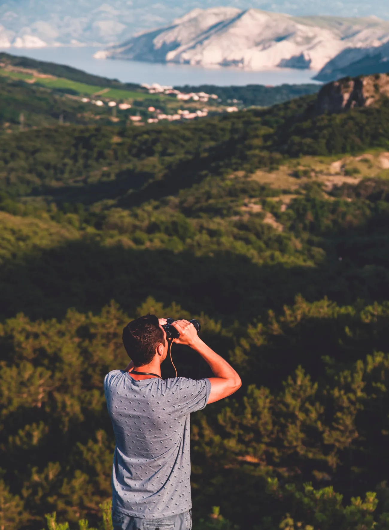 A man standing on a wooded hillside looking through binoculars across a wide valley towards distant mountains and the coast.